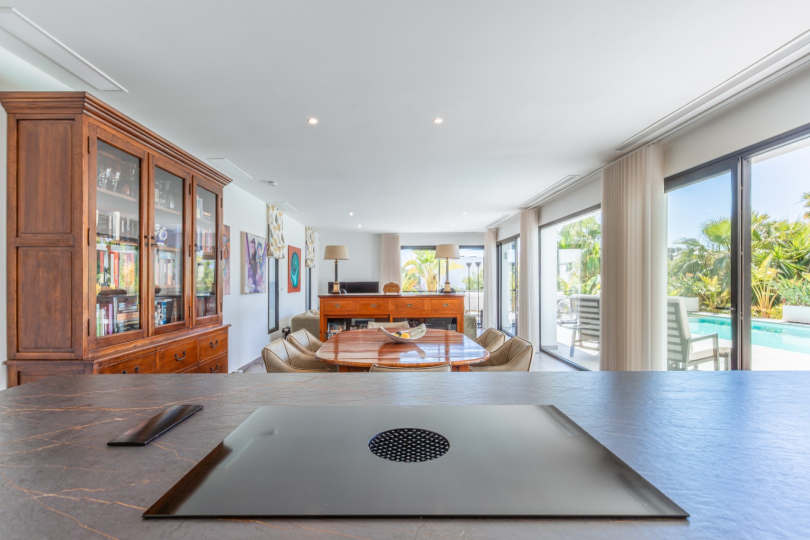 Vue depuis l'îlot de cuisine avec plaque à induction intégrée vers la salle à manger, soulignant le mobilier en bois et l'accès direct à la terrasse.