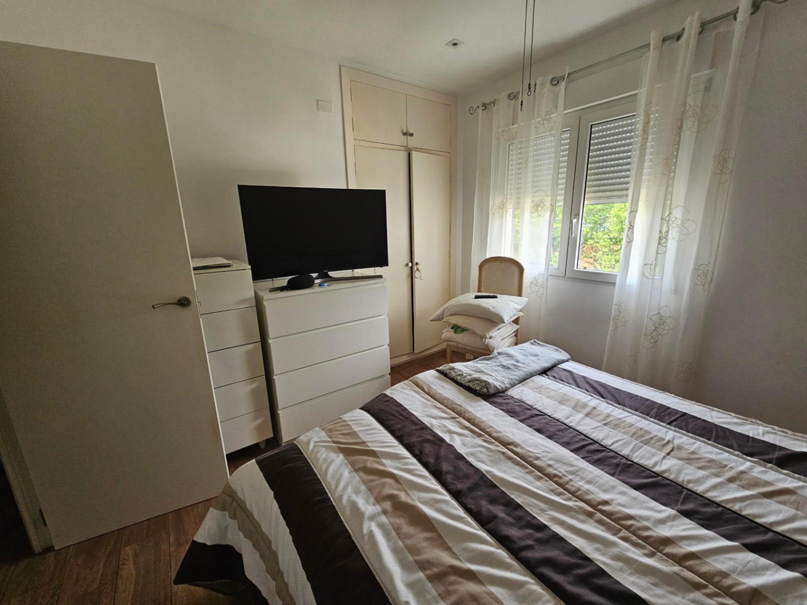 Chambre fonctionnelle avec placards intégrés, commodes blanches et une fenêtre avec volets roulants laissant entrer la lumière naturelle sur le sol stratifié.