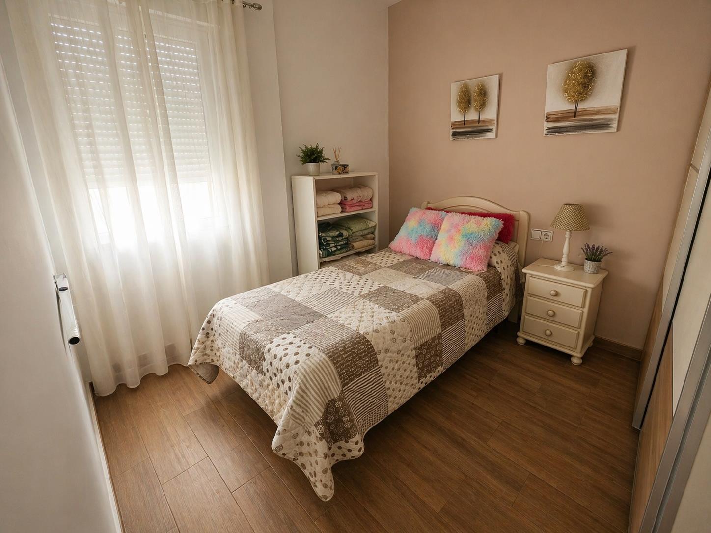 Chambre individuelle avec sol imitation bois, fenêtre équipée de stores et rideaux blancs, et un mur d'accent de couleur saumon orné de tableaux.
