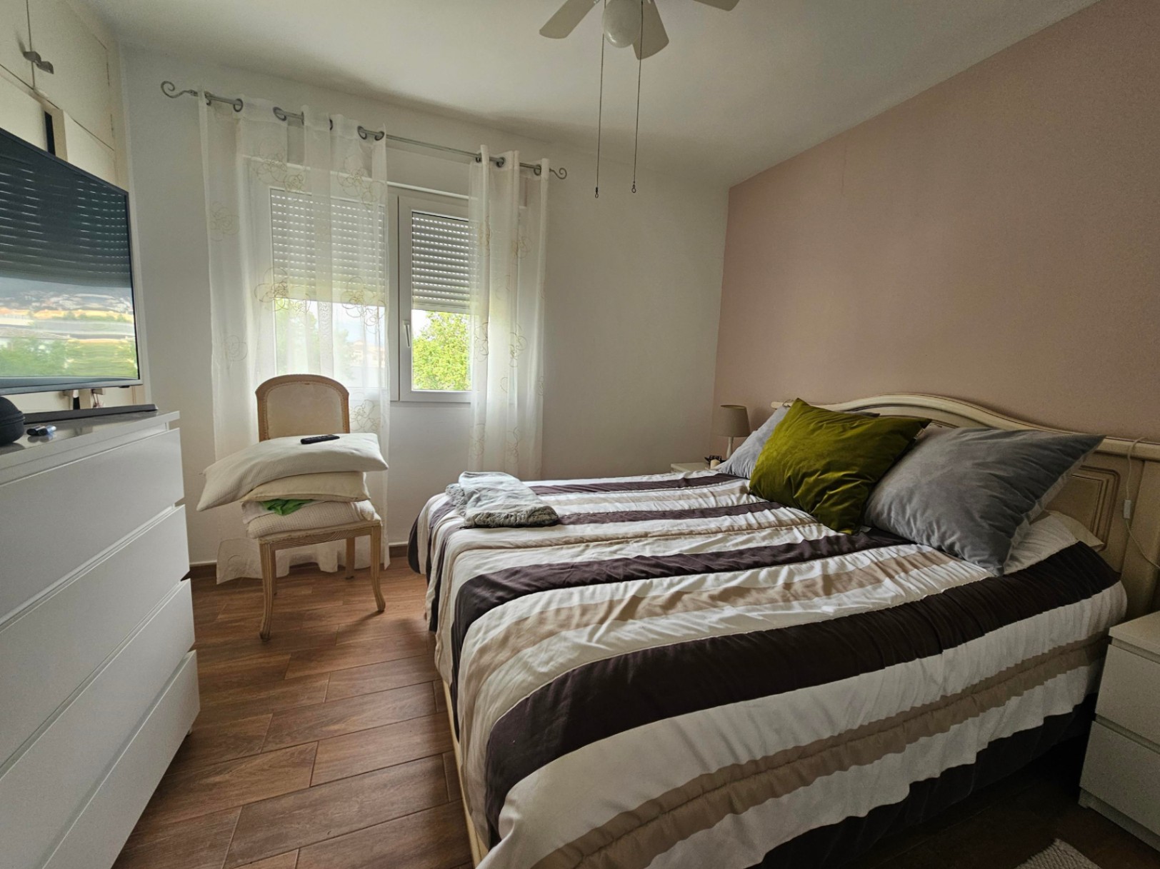 Chambre lumineuse avec sol en grès cérame imitation bois, mur d'accent saumon et ventilateur de plafond. La fenêtre est équipée de volets roulants et de rideaux légers.