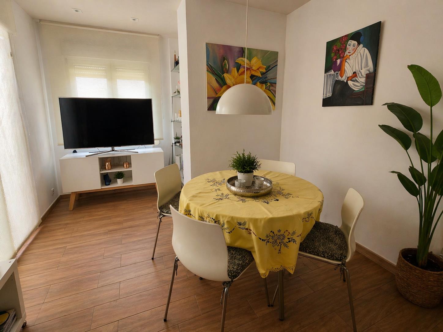 Coin repas avec table ronde, chaises blanches et luminaire suspendu, ouvert sur le salon avec sol en imitation bois et lumière naturelle.