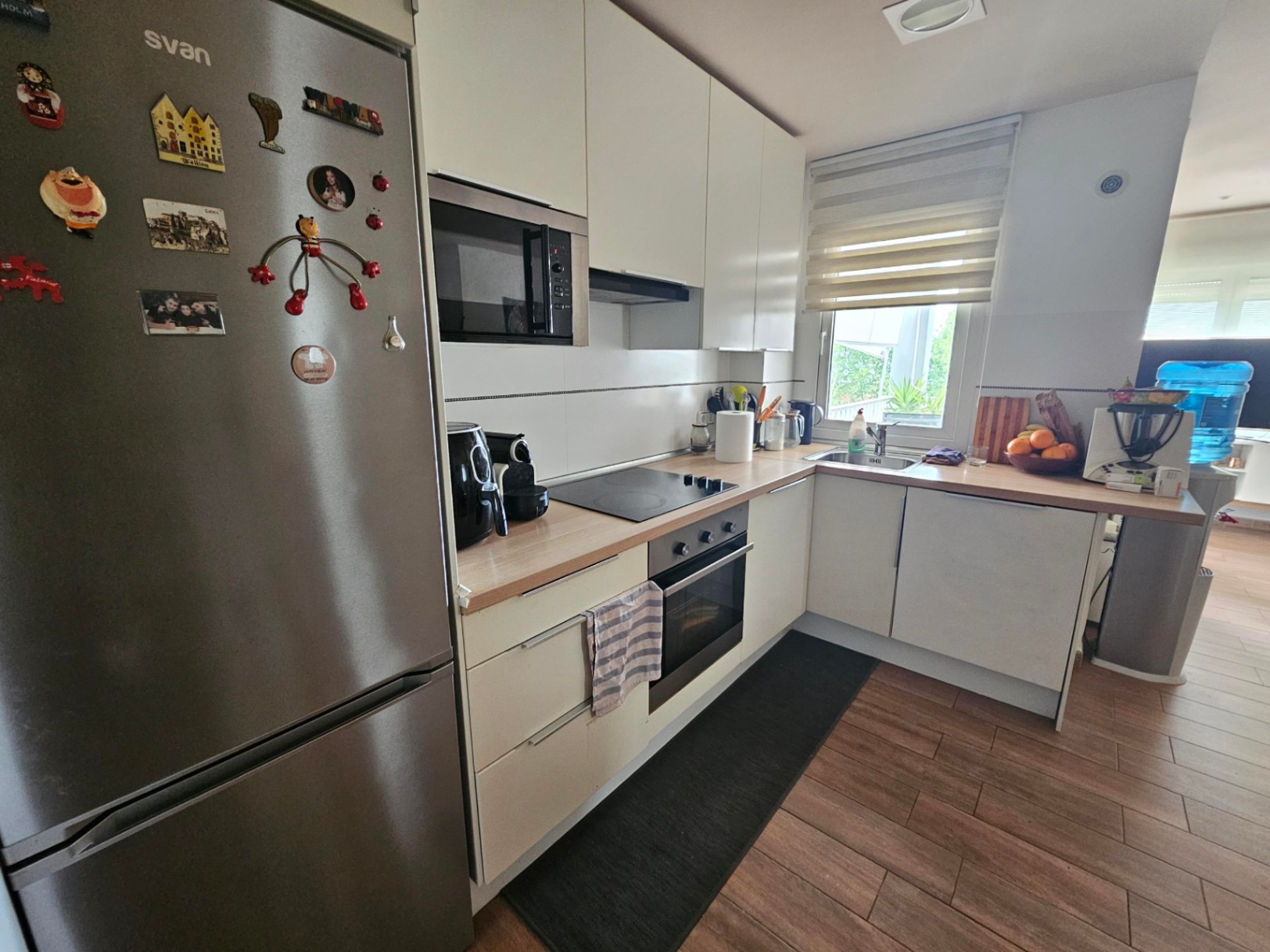Cuisine fonctionnelle en L avec plans de travail effet bois, électroménagers intégrés et fenêtre. Le sol en grès cérame imite la chaleur du bois.
