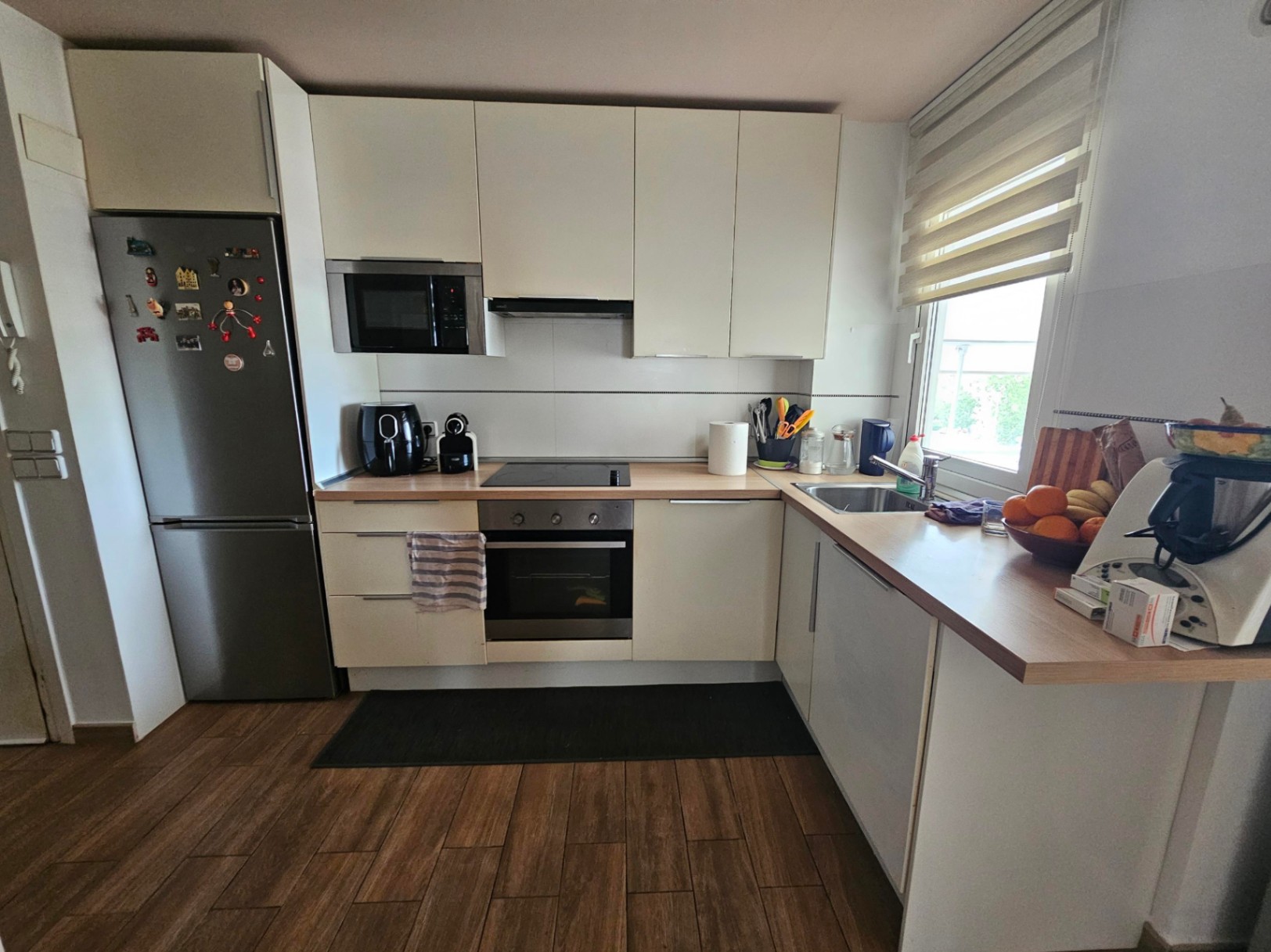 Cuisine moderne en L avec placards blancs, plan de travail en bois, appareils intégrés en acier inoxydable et sol imitation bois.
