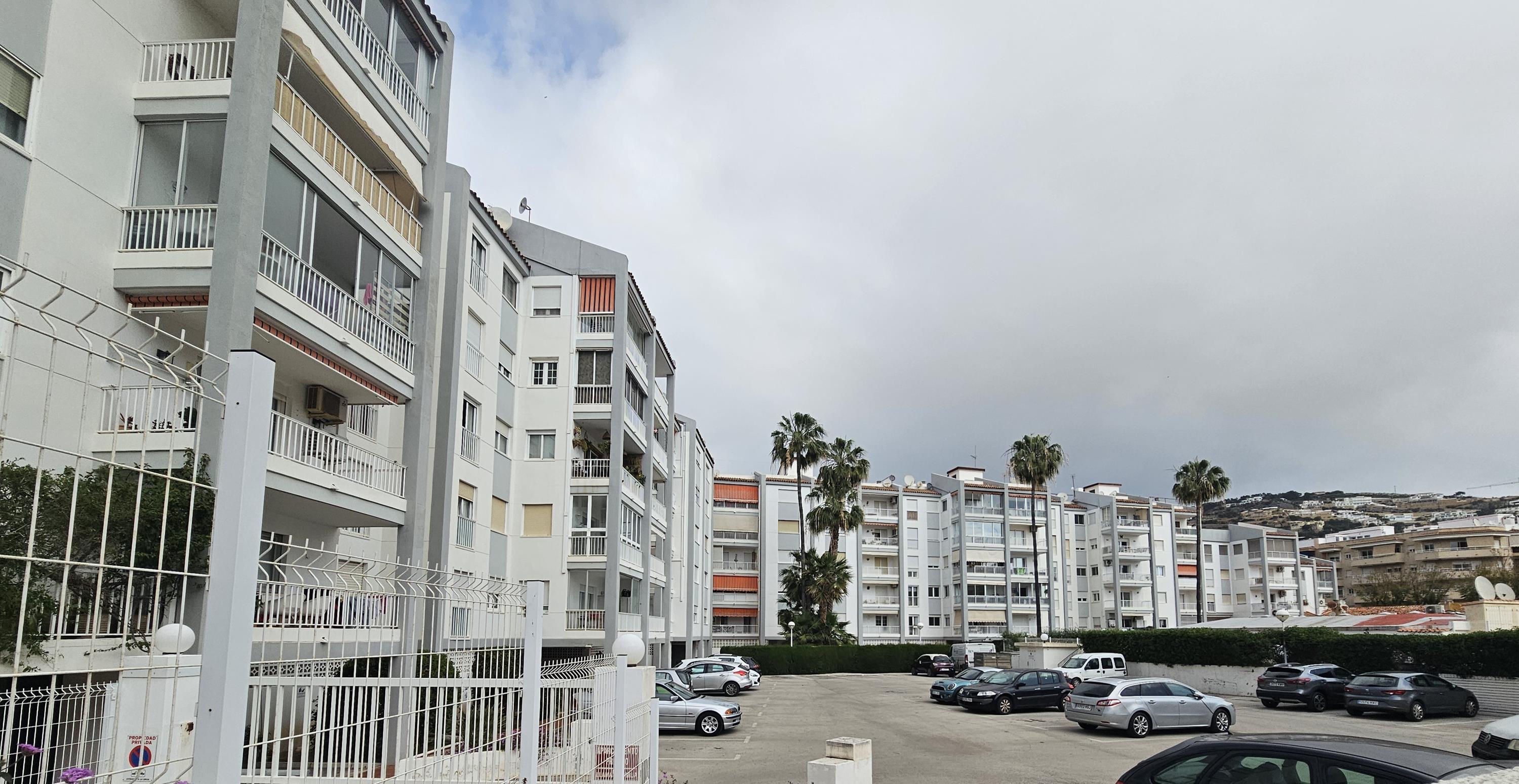 Façade du bâtiment résidentiel avec balcons et stores, comprenant un grand parking communautaire fermé et des espaces paysagers avec palmiers.