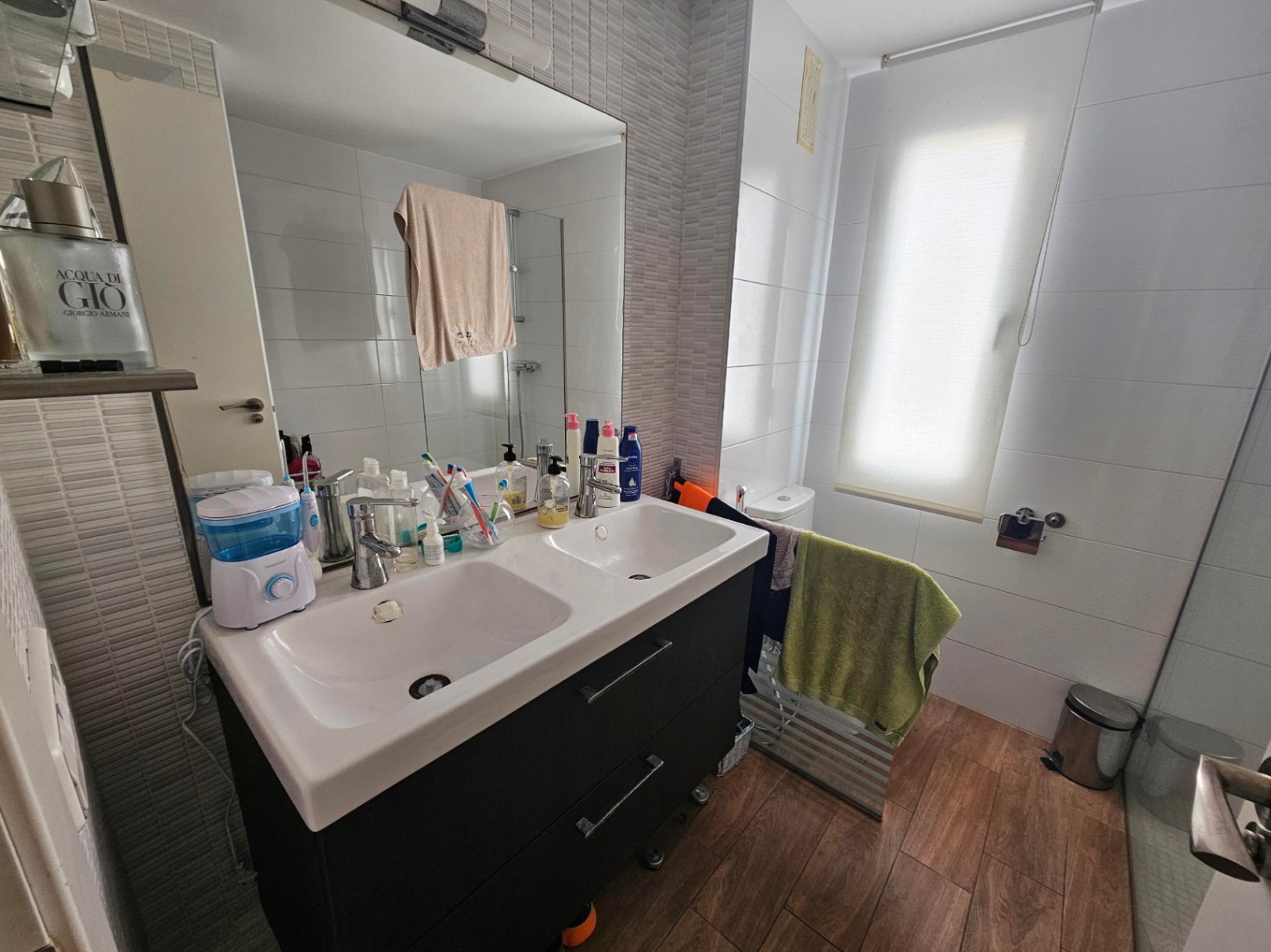 Salle de bain moderne avec meuble double vasque sombre, sol en grès cérame aspect bois et carrelage mural neutre bénéficiant d'une lumière naturelle.