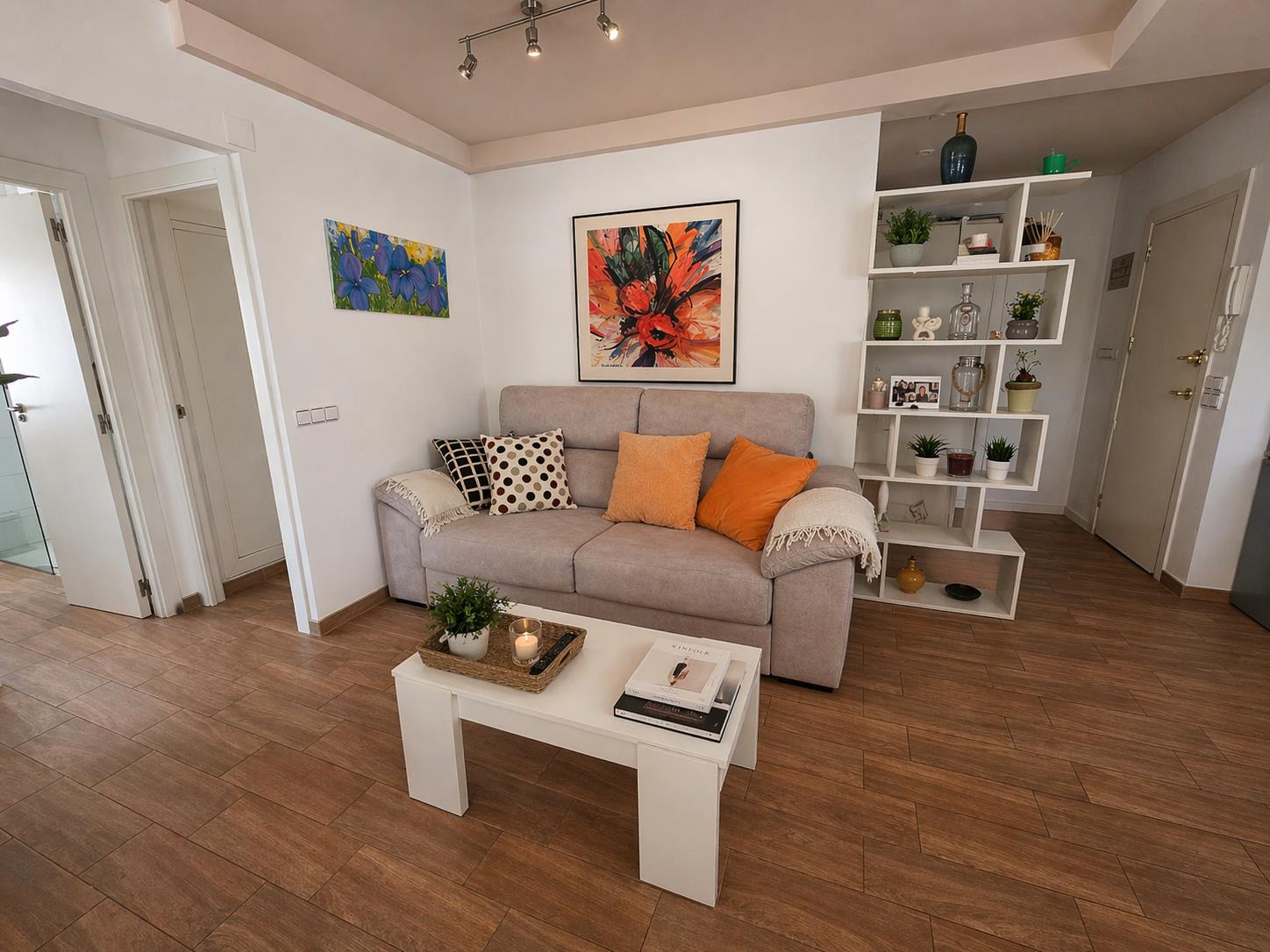 Salon chaleureux avec canapé en tissu, table basse blanche et sol en grès cérame imitation bois. Une étagère géométrique sépare élégamment l'espace de l'entrée.
