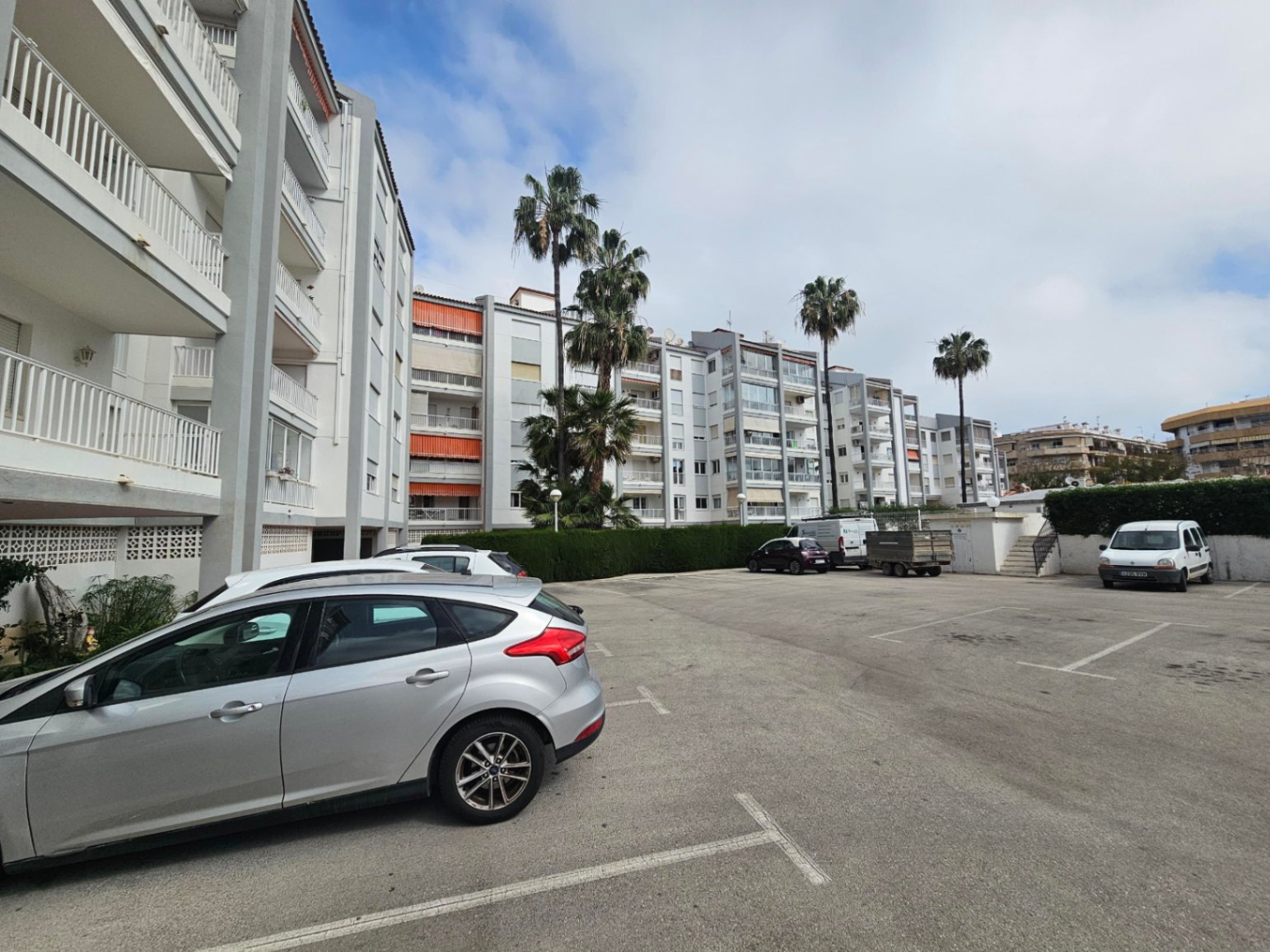 Zone de parking communautaire extérieur avec places délimitées, entourée de haies entretenues et de palmiers face à la façade du bâtiment.
