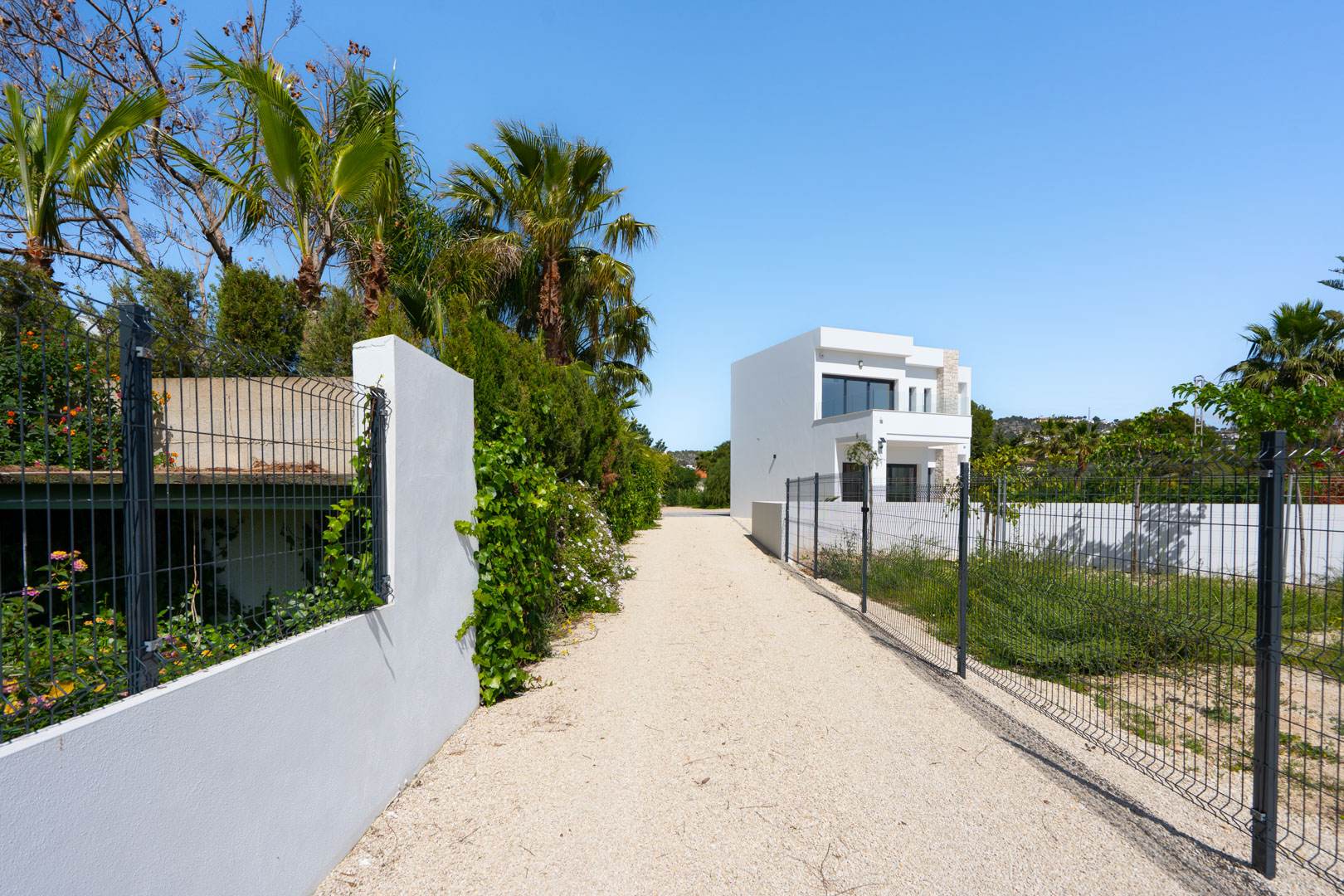 Allée d'accès en gravier menant à la villa moderne, bordée de clôtures, de palmiers méditerranéens et d'un vaste terrain plat.