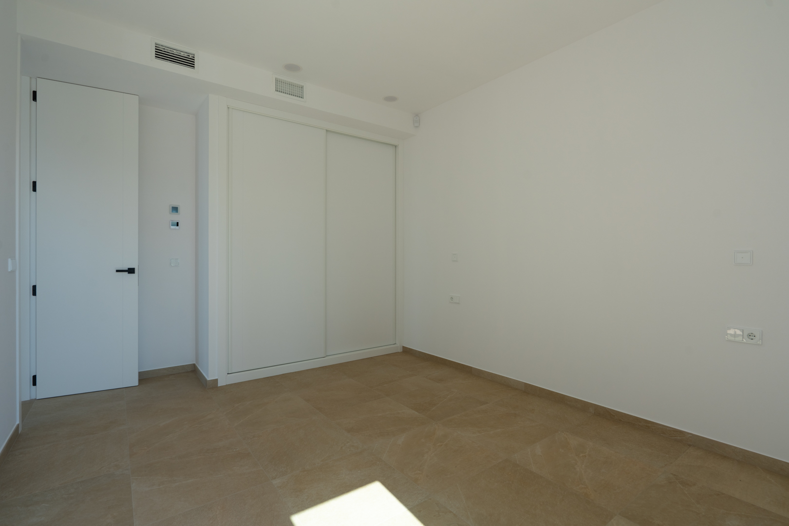 Chambre lumineuse avec sol en grès cérame aux tons terre, placard encastré à portes coulissantes blanches et climatisation par conduits intégrée.