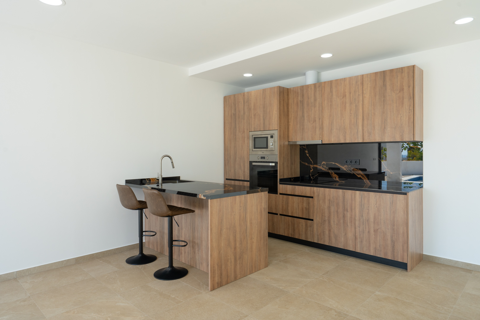 Cuisine de designer avec façades en bois, plan de travail en pierre noire veinée et îlot central avec tabourets. Équipée d'appareils intégrés et d'un éclairage encastré.