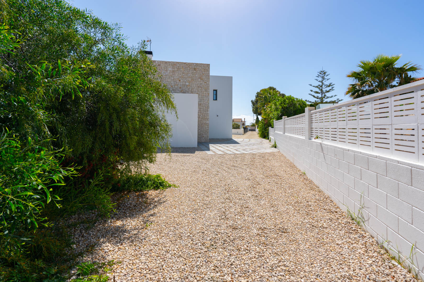 Entrée latérale avec allée en gravier, mur d'enceinte blanc et façade alliant pierre naturelle et finitions blanches modernes.