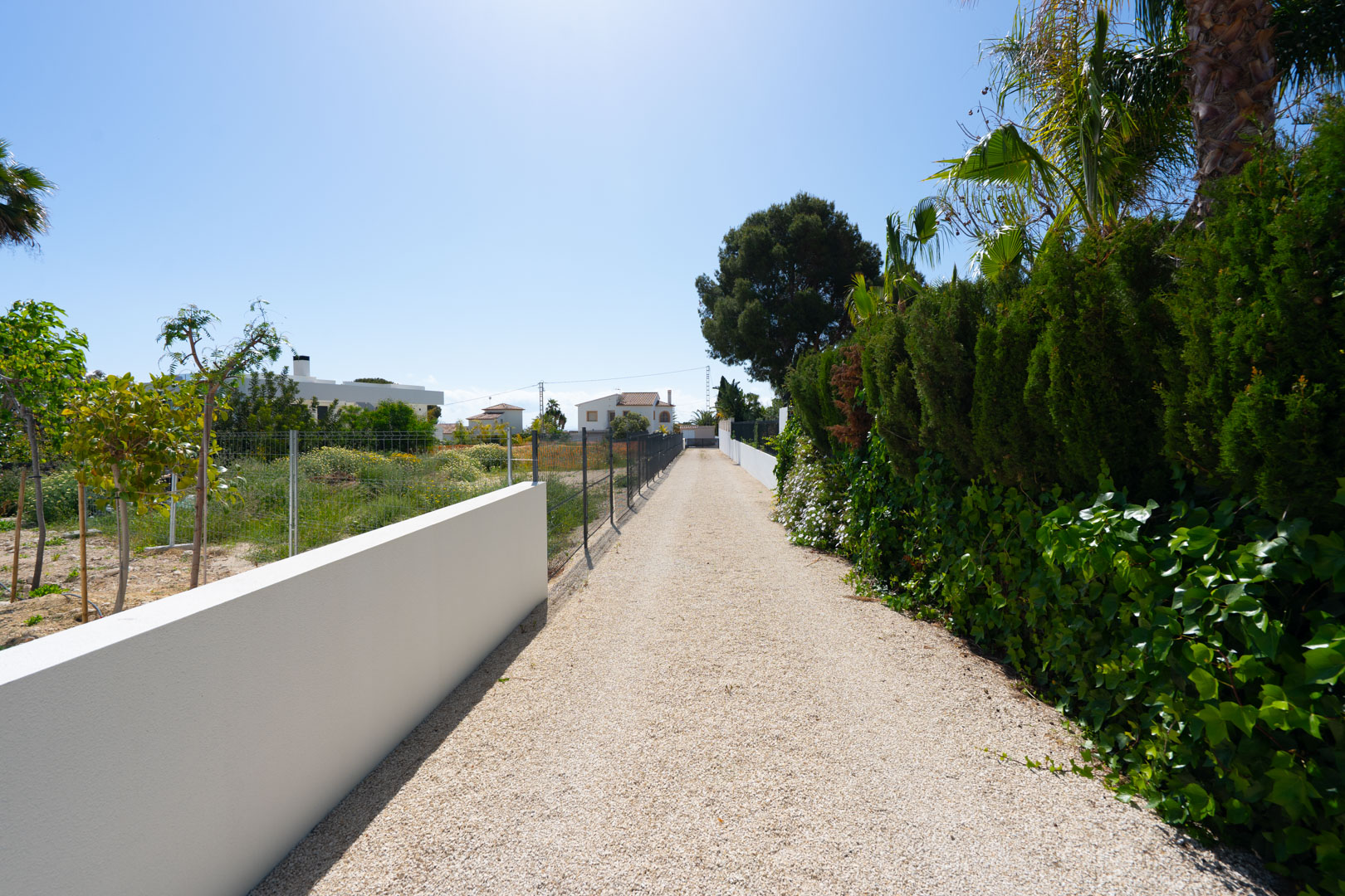 Longue allée en gravier bordée d'un mur blanc épuré et d'une haie verdoyante, illustrant l'étendue du terrain plat et privé de la propriété.