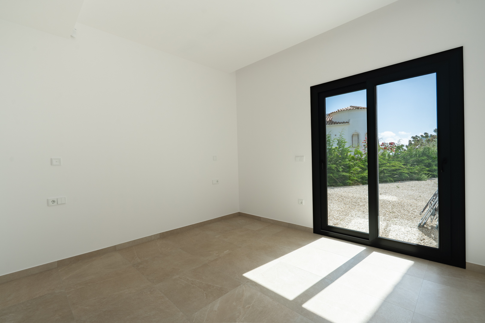 Pièce lumineuse avec sol en grès cérame et grande baie vitrée coulissante en aluminium noir offrant un accès direct aux espaces extérieurs.