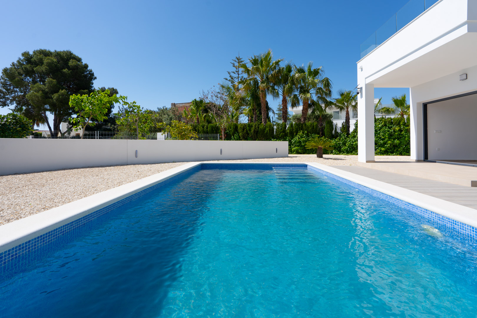 Piscine rectangulaire en mosaïque bleue entourée de gravier blanc et de murs d'enceinte modernes, avec des palmiers et une végétation soignée.