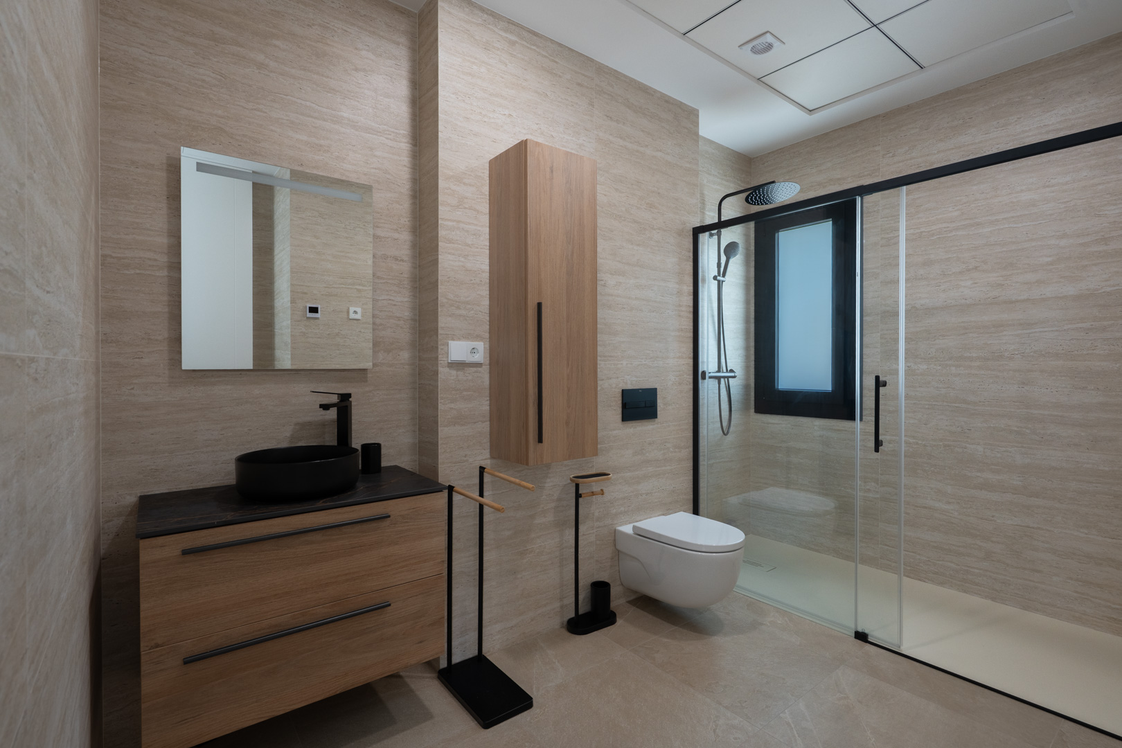 Salle de bain design avec carrelage effet travertin, meuble vasque en bois avec vasque noire et douche à l'italienne dotée de finitions noir mat.