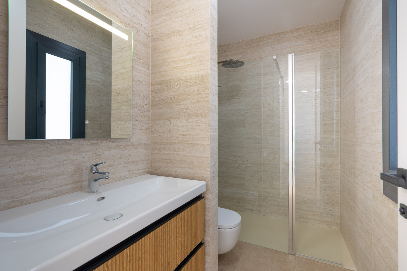 Salle de bains design avec carrelage en grès cérame effet pierre, meuble vasque en bois rainuré et douche à l'italienne avec paroi vitrée et miroir à éclairage LED intégré.