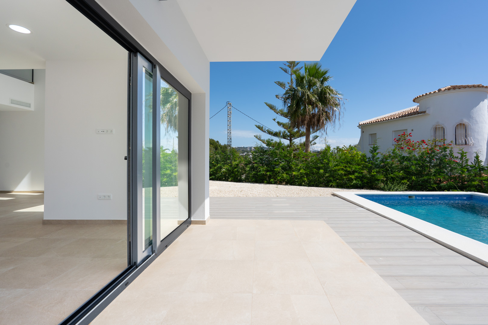 Terrasse couverte avec carrelage en grès cérame, ouvrant sur une piscine carrelée de bleu et un jardin paysager avec des palmiers sous un ciel dégagé.