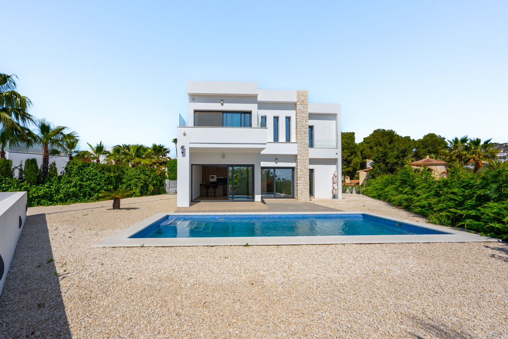 Villa moderne à deux étages avec façade blanche, mur en pierre naturelle, piscine rectangulaire et aménagement paysager en gravier entouré de végétation.