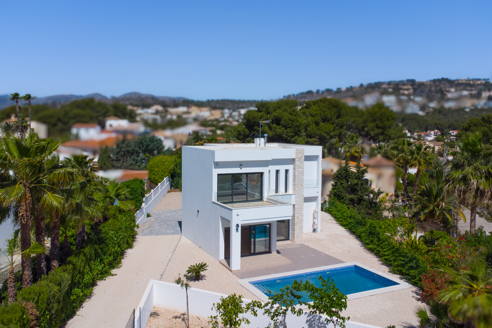 Vue extérieure de la villa moderne avec façade blanche, accents en pierre naturelle, piscine rectangulaire et terrasses avec garde-corps en verre sur un vaste terrain plat.