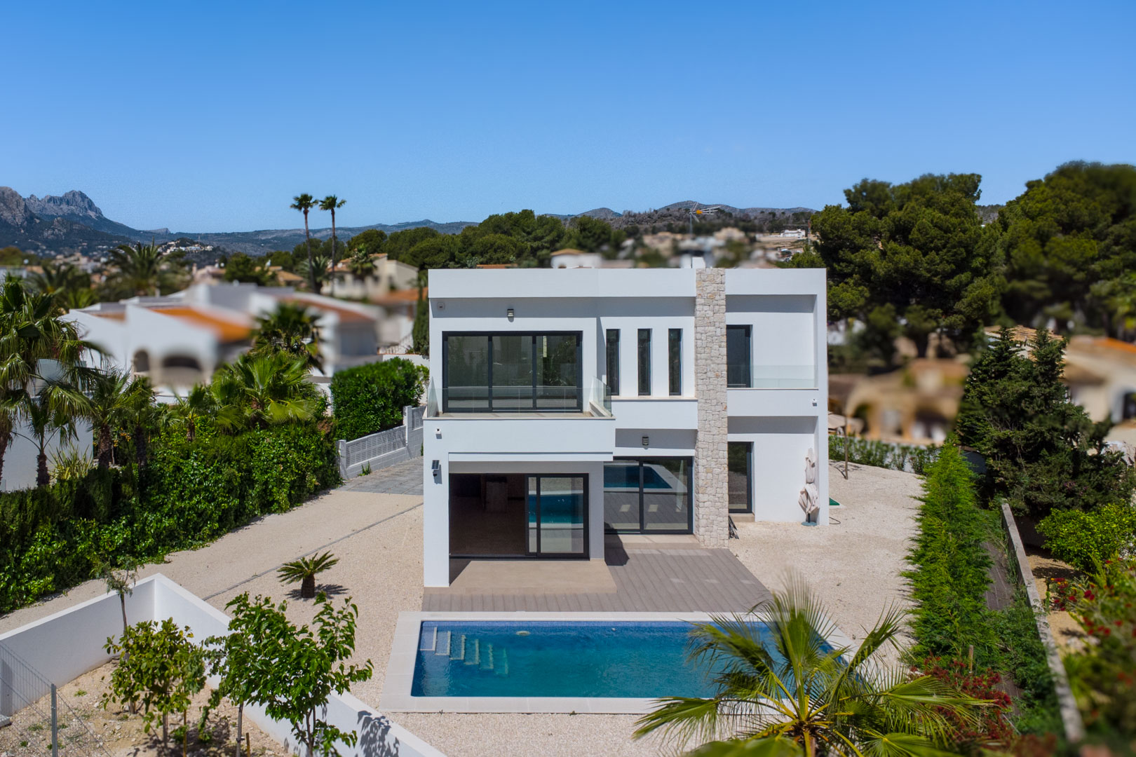Vue extérieure de la villa moderne avec façade blanche, pilier en pierre naturelle, piscine rectangulaire et terrasses avec garde-corps en verre.