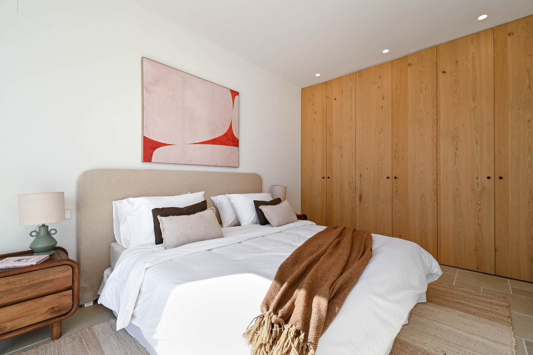 Chambre avec de grands placards intégrés en bois naturel, une tête de lit rembourrée beige et un éclairage encastré. Le sol est revêtu de carrelage clair avec un tapis texturé.