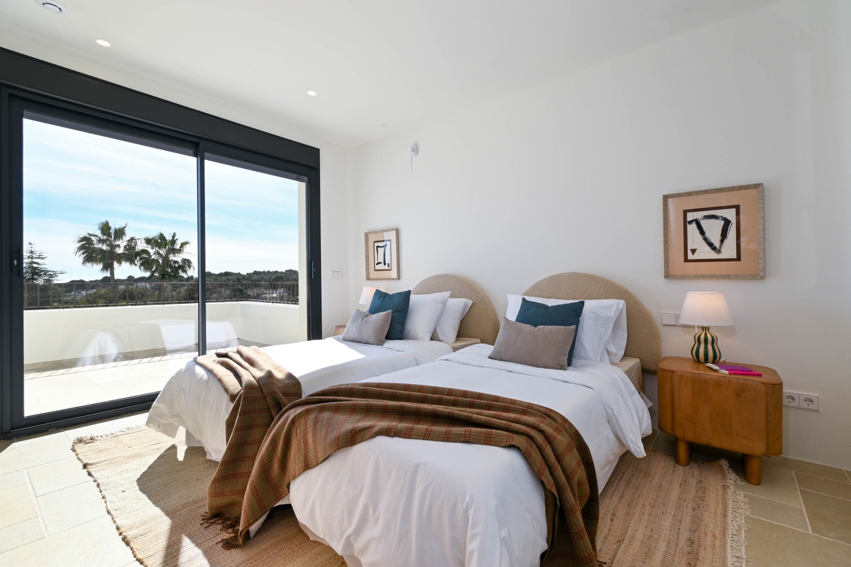 Chambre lumineuse avec deux lits simples, têtes de lit arrondies et accès direct à une terrasse offrant une vue dégagée sur les palmiers et la nature.