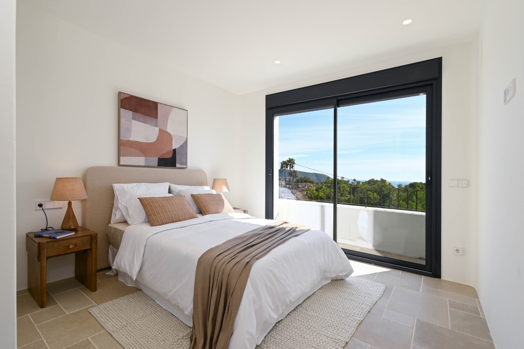 Chambre lumineuse avec sol en pierre naturelle et grandes baies vitrées coulissantes ouvrant sur une terrasse privée offrant une vue dégagée sur les environs.