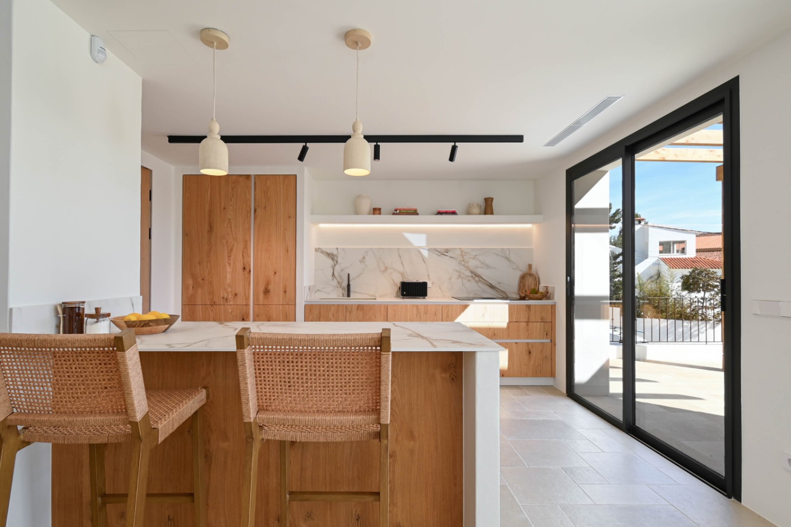 Cuisine à aire ouverte avec îlot en bois, plans de travail effet marbre et tabourets en osier, ouvrant sur la terrasse par de grandes baies vitrées noires.
