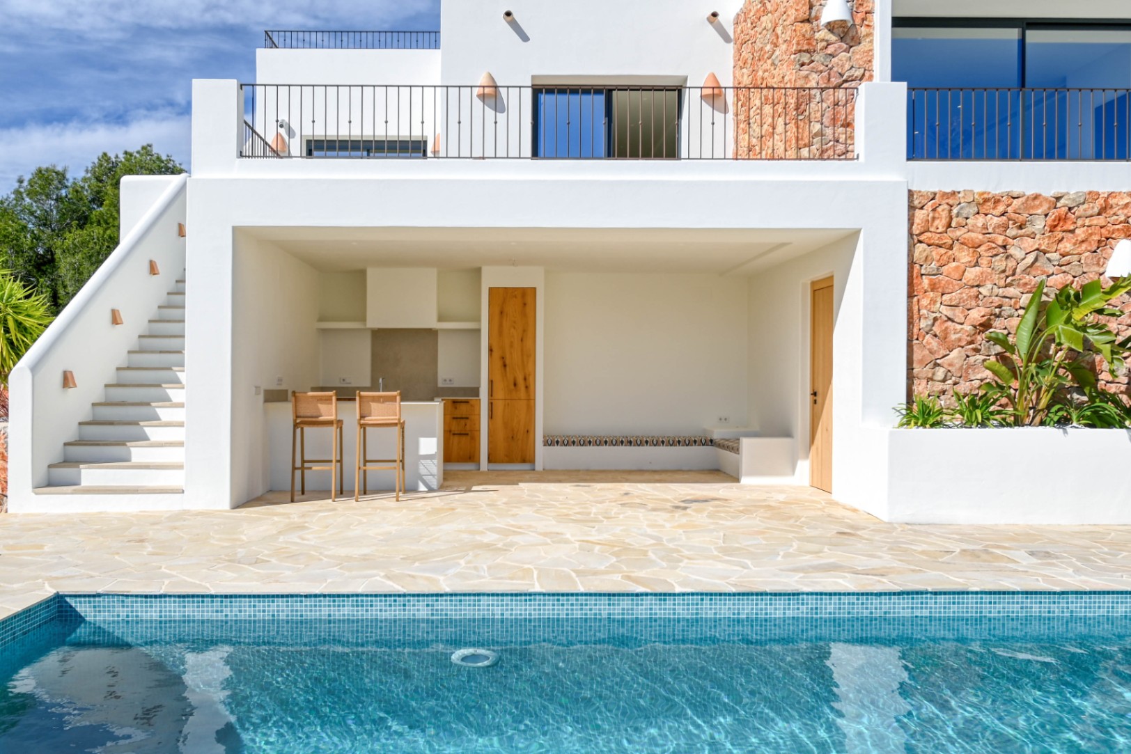 Cuisine d'été et espace détente au bord de la piscine, avec murs en pierre naturelle, menuiseries en bois et terrasse en dalles de pierre sous une façade blanche.