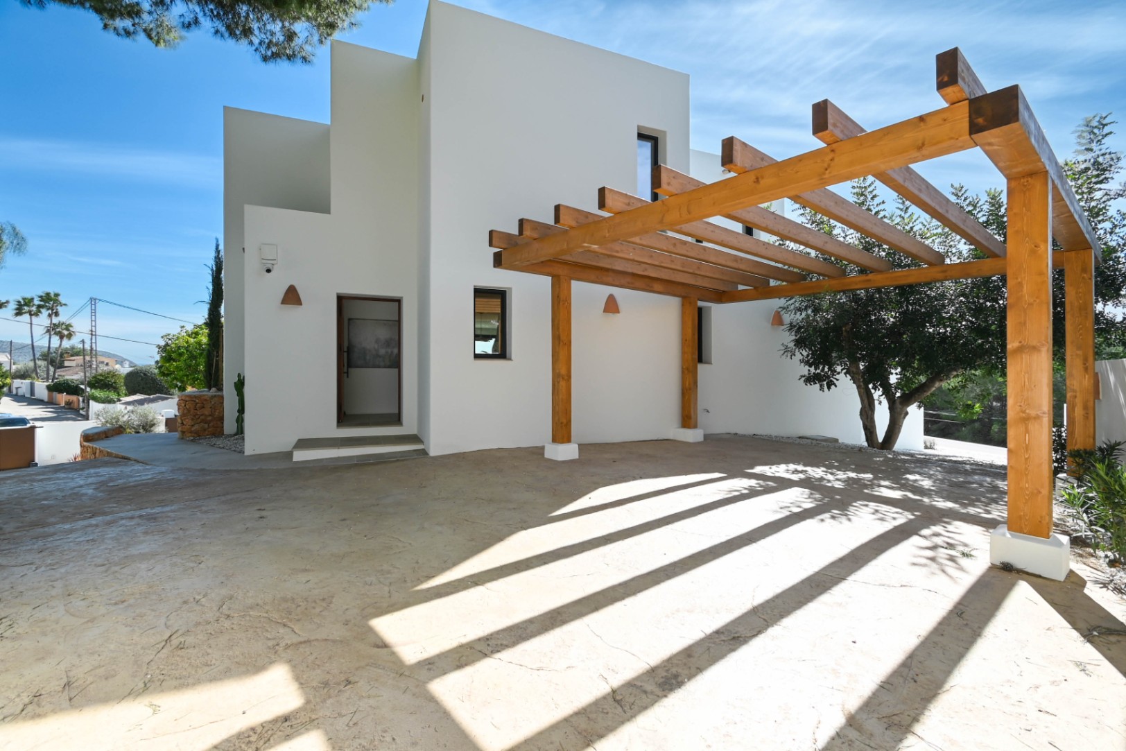 Façade blanche minimaliste avec une grande pergola en poutres de bois naturel sur une zone de stationnement pavée. Comprend des appliques murales en céramique.