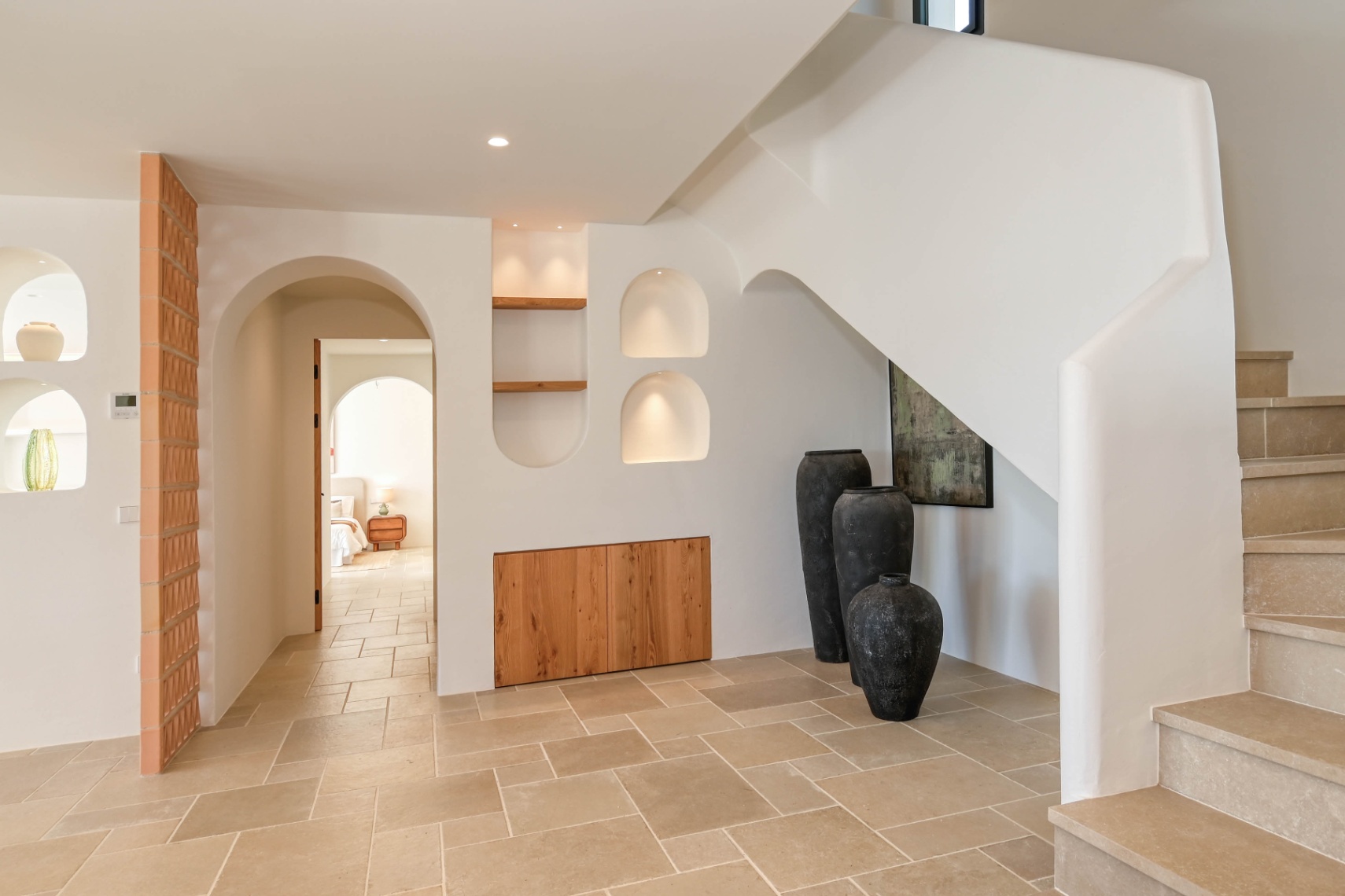 Hall d'entrée méditerranéen avec escalier en pierre, niches murales éclairées et placards en bois intégrés. Le sol en pierre naturelle s'harmonise avec les arches épurées.