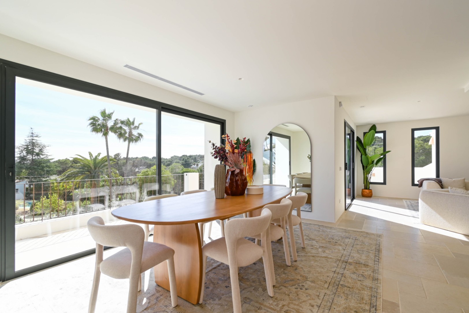 Salle à manger lumineuse avec table ovale en bois et grandes baies vitrées coulissantes ouvrant sur une terrasse avec vue sur les palmiers et la végétation.