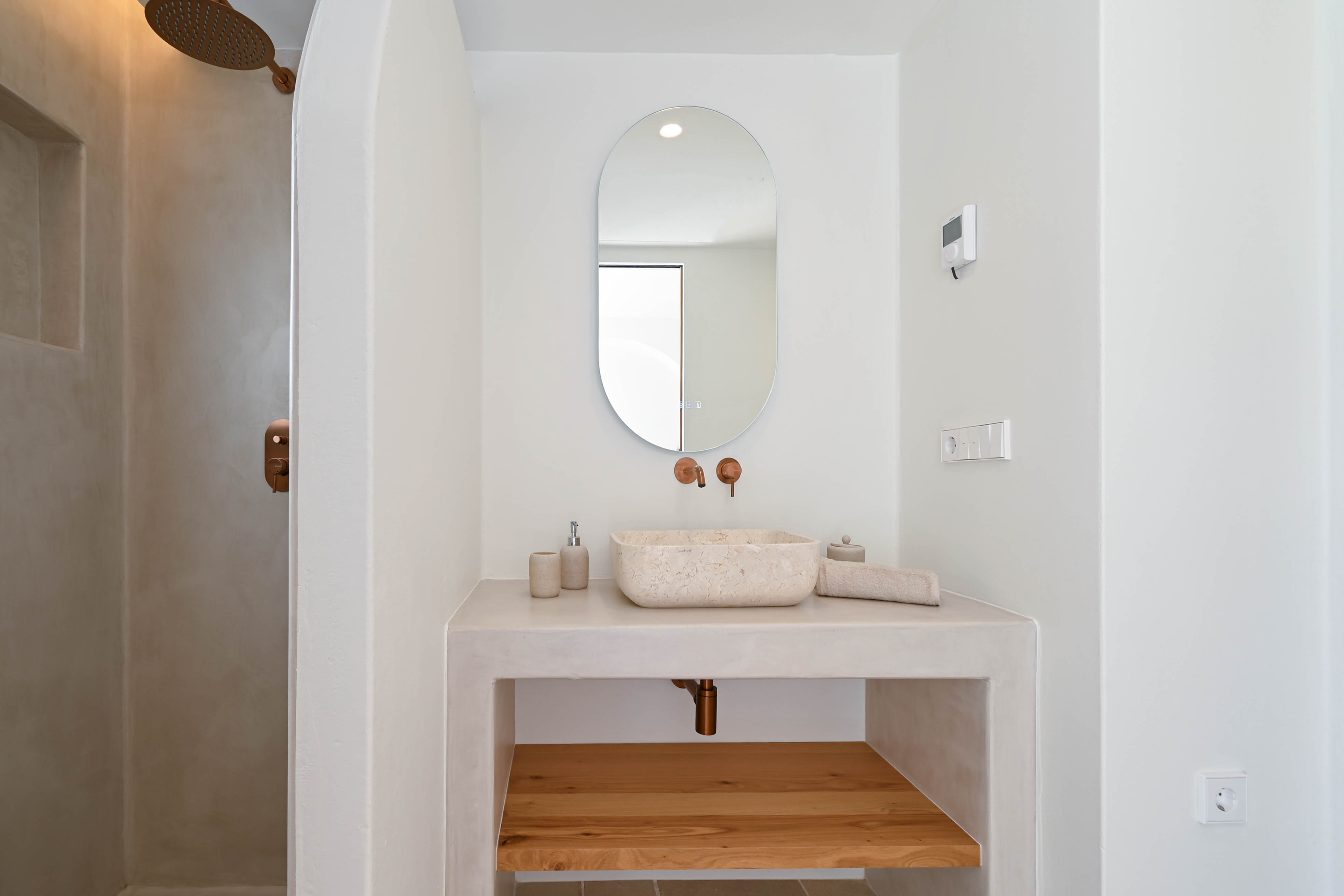 Salle de bain moderne avec finitions en microciment, vasque en pierre naturelle et robinetterie en cuivre. Elle dispose d'une étagère en bois et d'une douche à l'italienne.