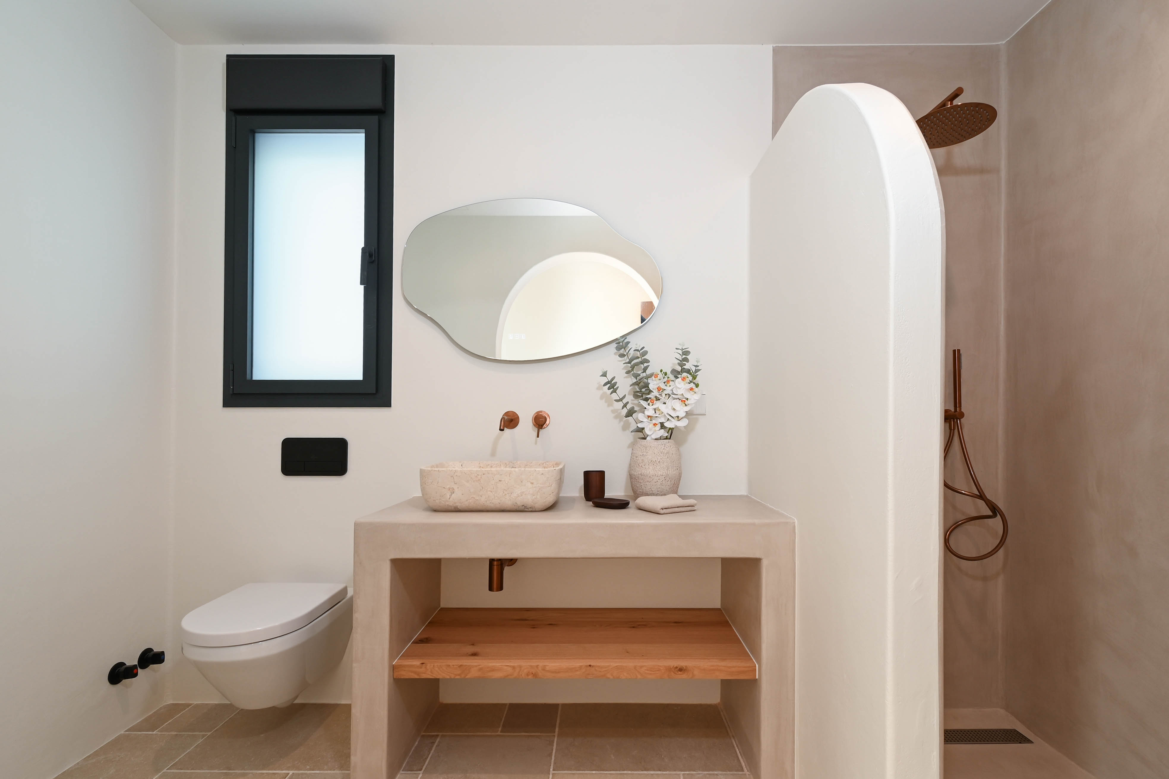 Salle de bain moderne avec plan en microciment, vasque en pierre naturelle et robinetterie en cuivre. Elle dispose d'un miroir organique, WC suspendu et douche avec paroi incurvée.
