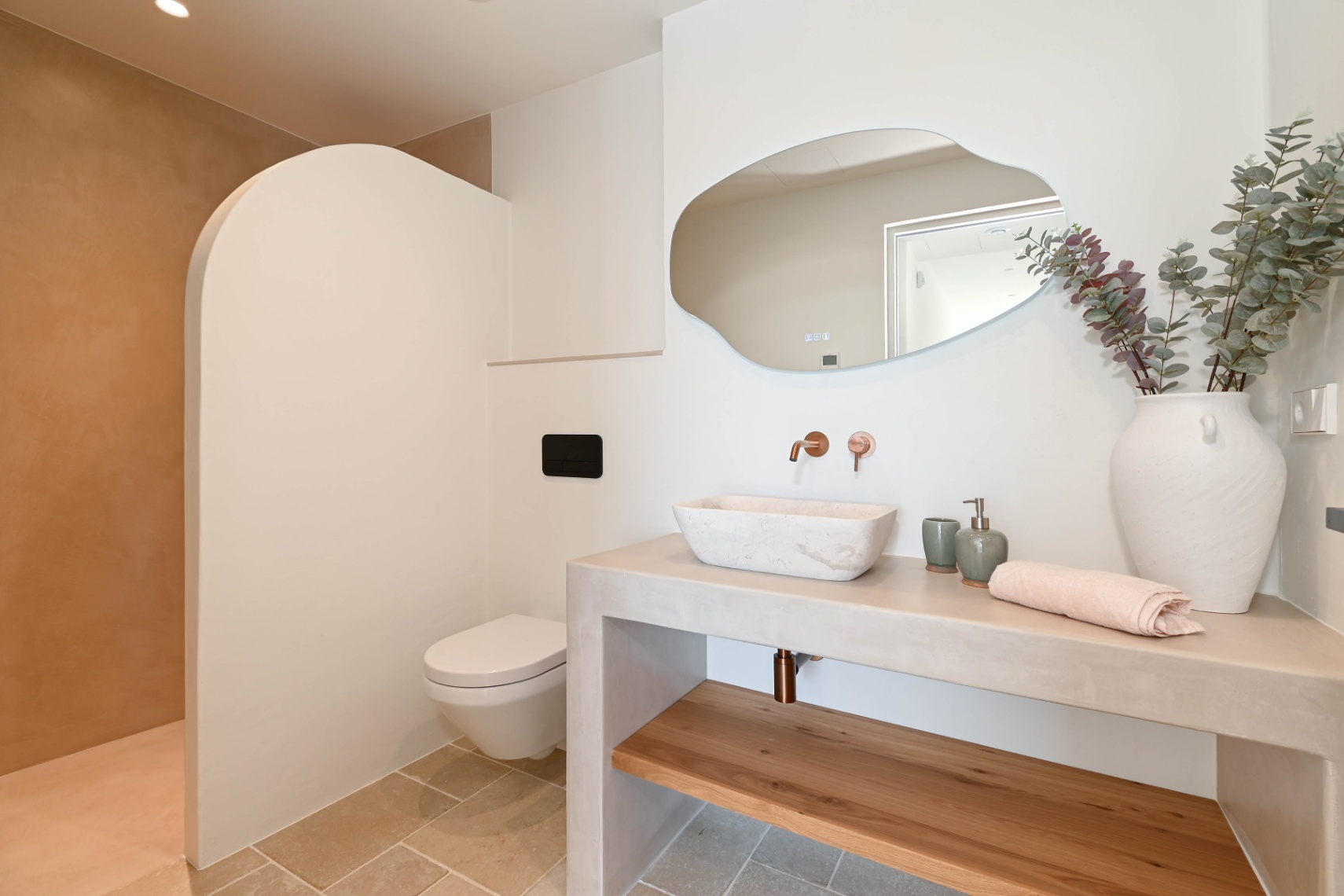 Salle de bain moderne avec vasque en pierre sur plan en microciment, robinetterie encastrée en cuivre, miroir organique et douche avec paroi incurvée élégante.