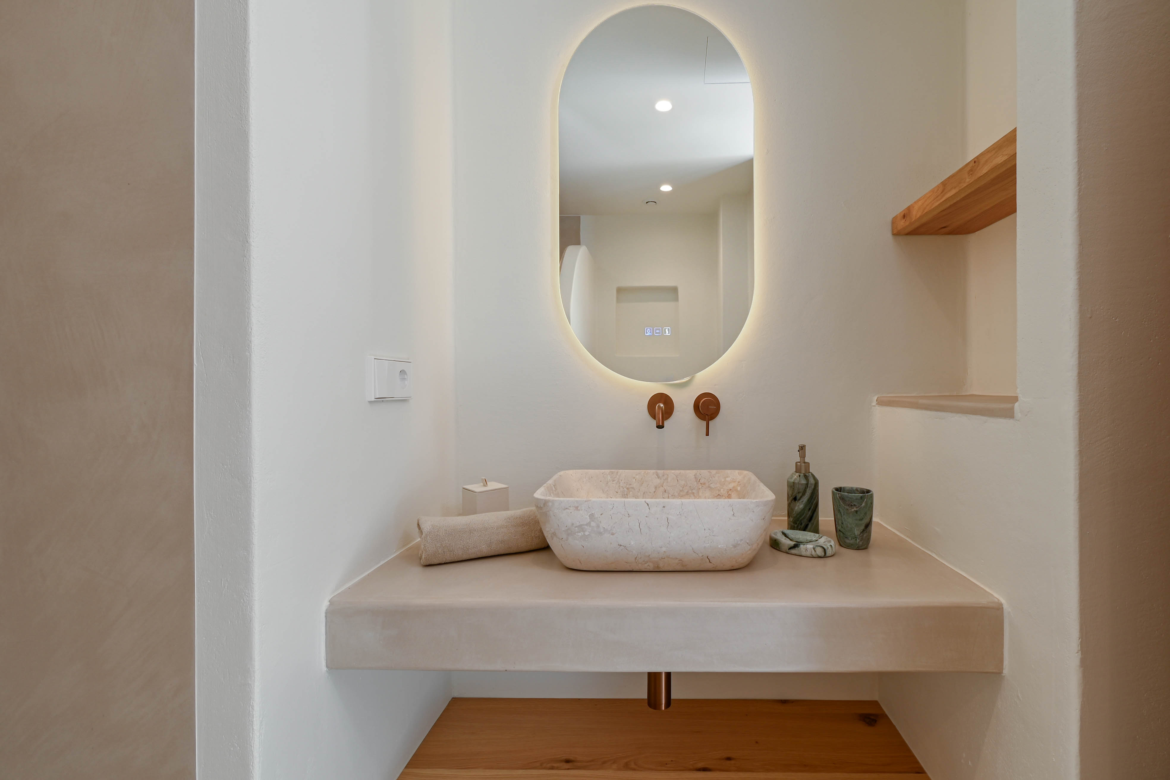 Salle de bains design avec vasque en pierre naturelle sur plan en microciment, robinetterie encastrée en cuivre et miroir ovale rétroéclairé.