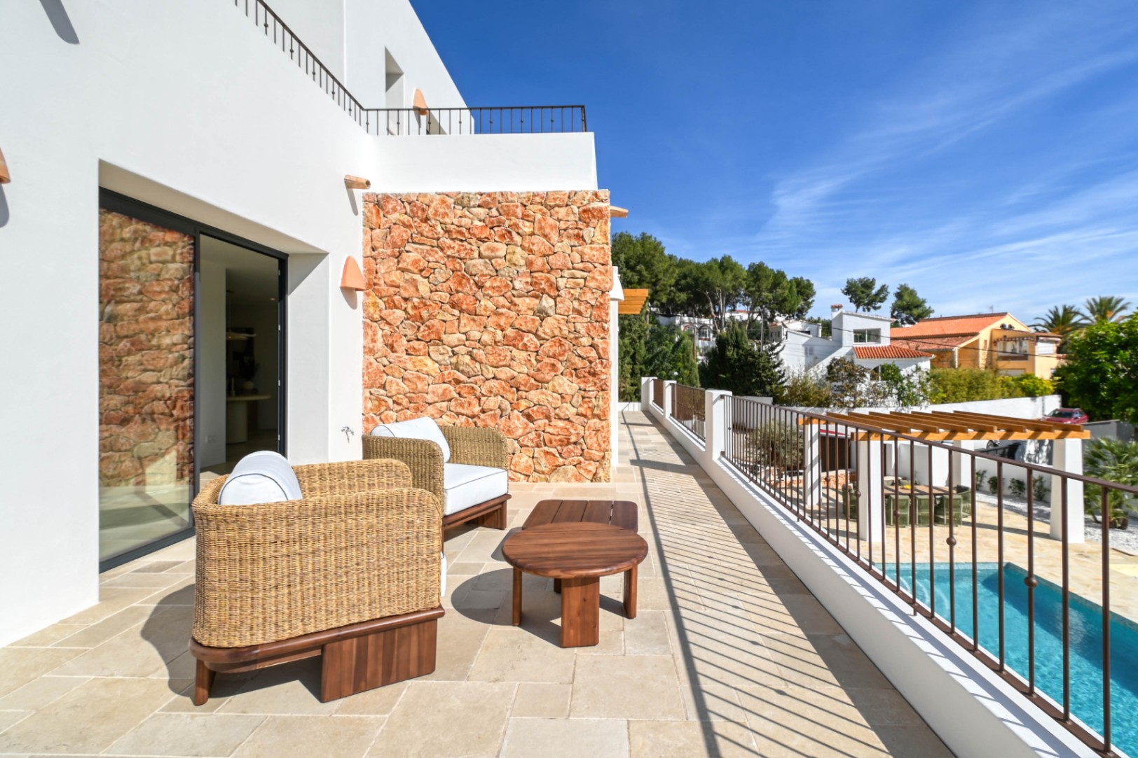 Terrasse supérieure dotée d'un sol en pierre, de fauteuils en osier et d'un mur en pierre naturelle, surplombant la piscine et l'espace repas sous pergola.