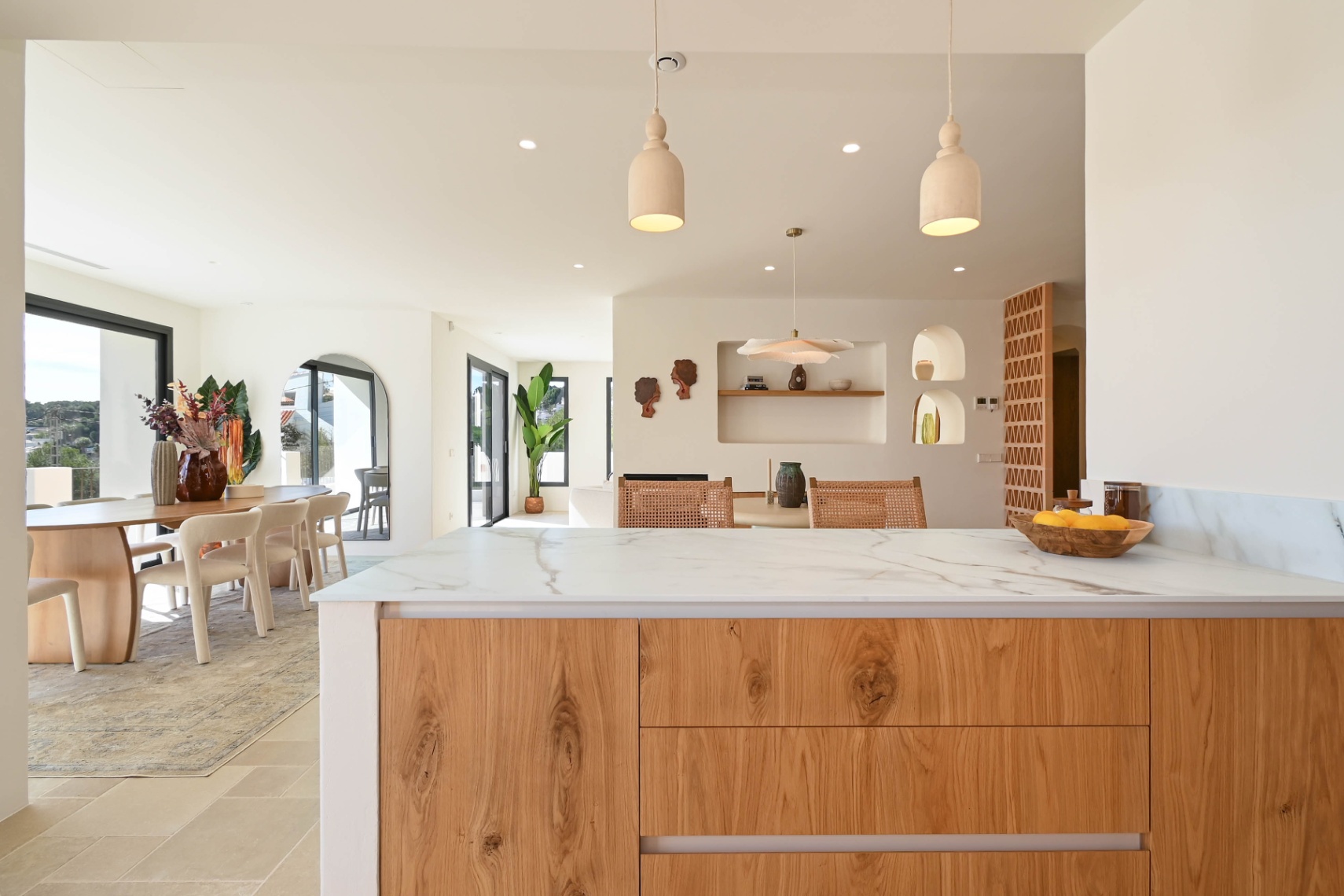 Vue depuis l'îlot de cuisine en pierre veinée vers la salle à manger, soulignant les niches murales décoratives, les finitions en bois et les grandes ouvertures vitrées.