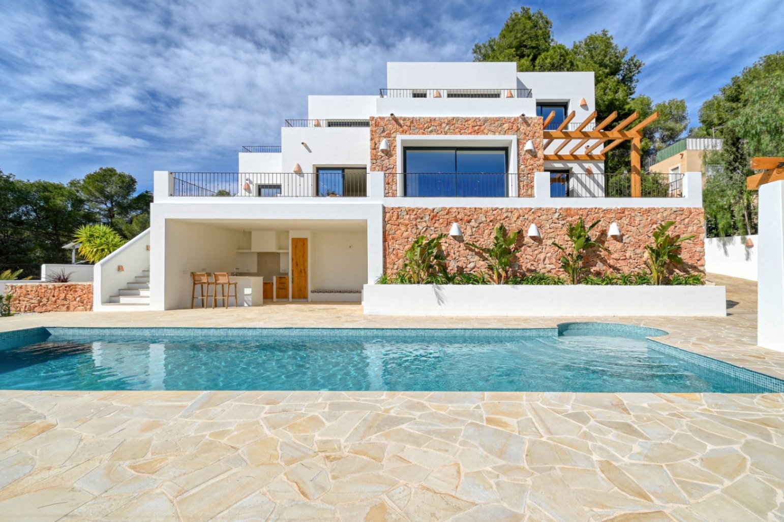 Vue extérieure avec piscine en mosaïque bleue, terrasse en pierre naturelle et façade à plusieurs niveaux alliant murs en pierre sèche, cuisine d'été et pergola en bois.
