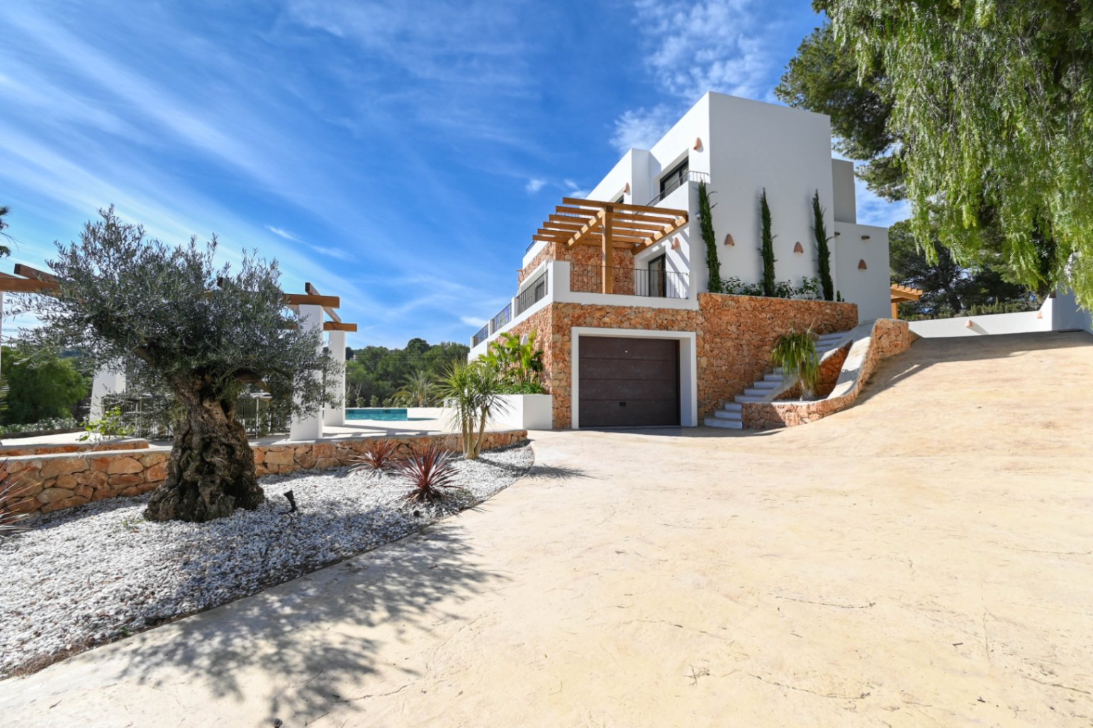 Vue extérieure de la villa avec façade en pierre naturelle, garage intégré et allée pavée agrémentée d'un olivier et d'un jardin méditerranéen.
