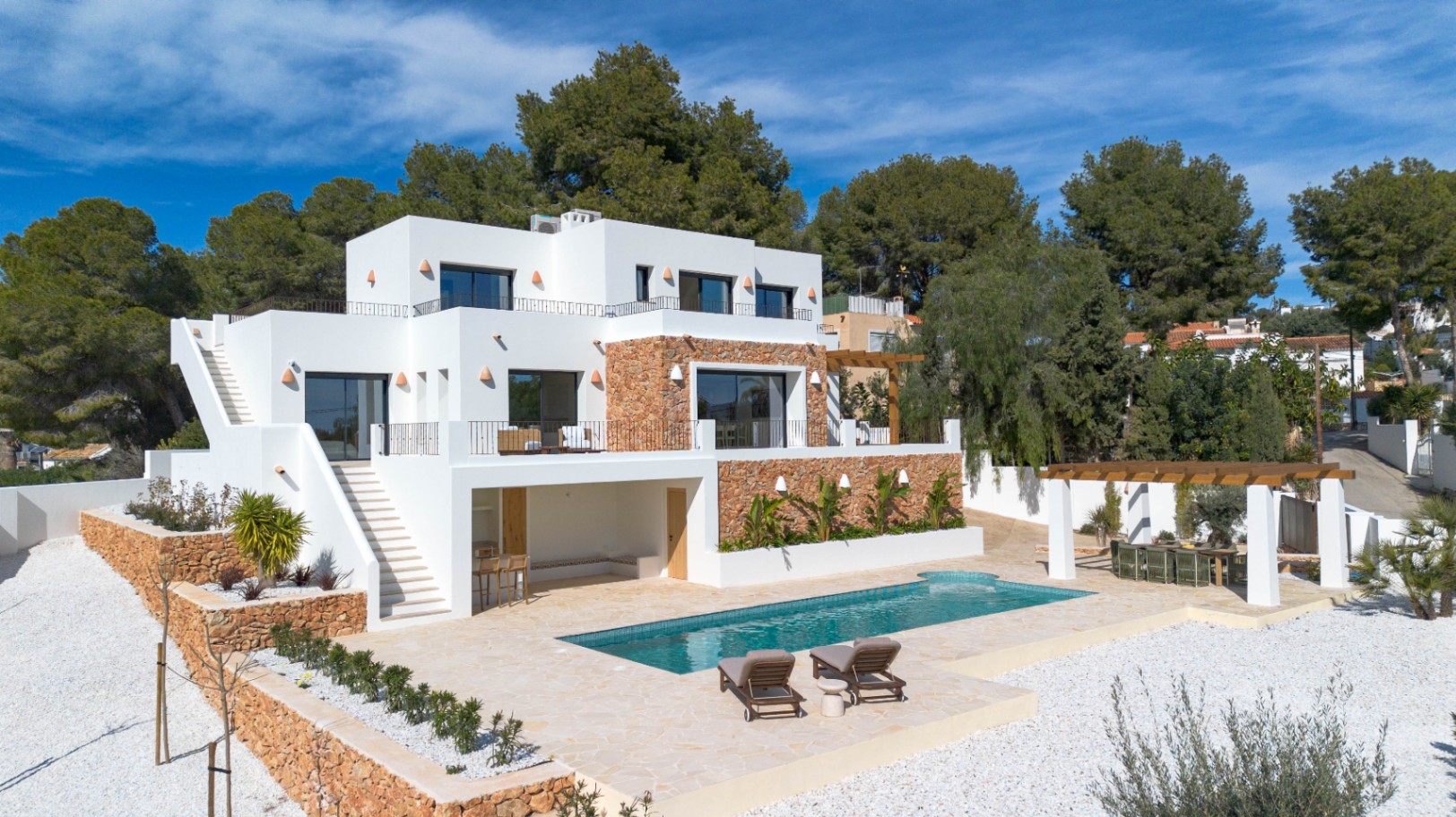 Vue extérieure de la villa moderne avec façades blanches, murs en pierre naturelle, piscine rectangulaire et terrasses à plusieurs niveaux entourées de végétation.
