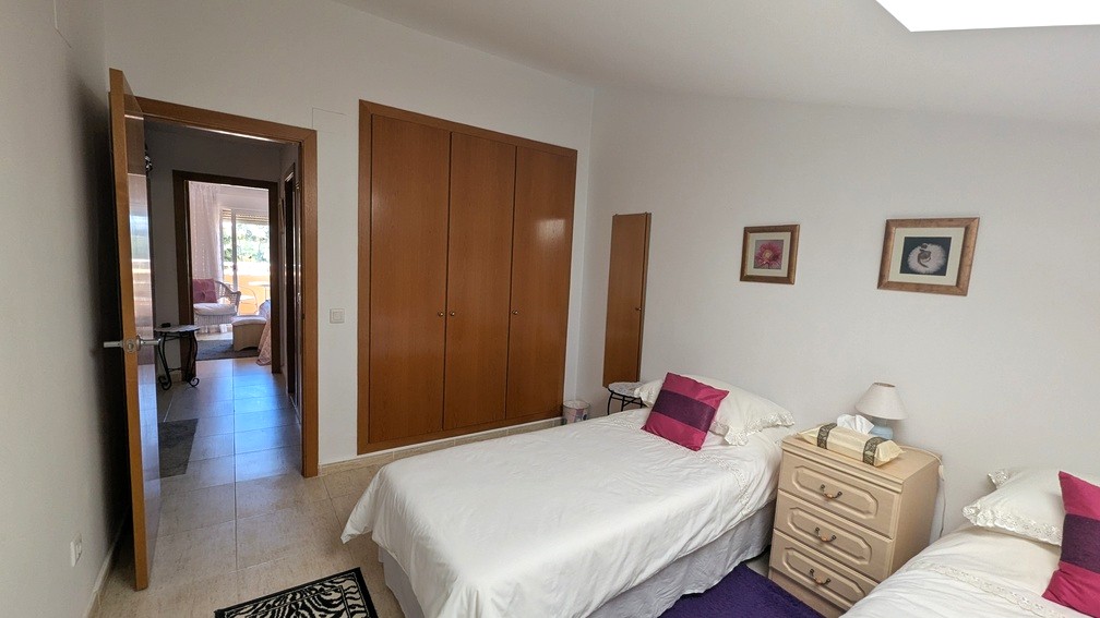 Chambre à deux lits avec placards intégrés en bois et sol carrelé. La porte ouverte donne sur le couloir menant à une pièce lumineuse avec accès à la terrasse.