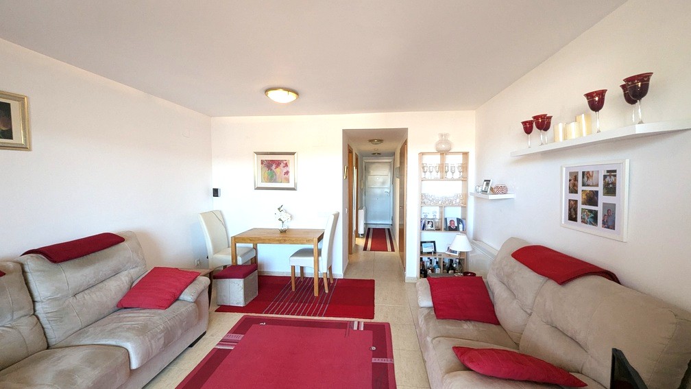Espace salon-salle à manger avec canapés beiges, touches rouges et table en bois. La pièce s'ouvre sur un couloir central avec carrelage clair et murs blancs.