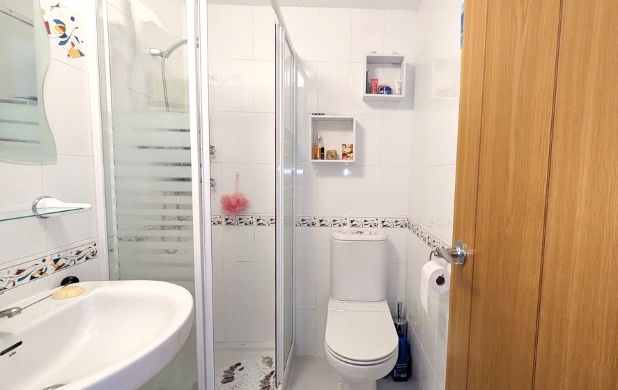 Salle de douche moderne avec murs carrelés blancs et frise décorative. Comprend une cabine de douche vitrée à bandes dépolies, un lavabo suspendu et des toilettes, avec deux étagères murales. Salle de douche moderne avec murs carrelés blancs et frise décorative. Comprend une cabine de douche vitrée à bandes dépolies, un lavabo suspendu et des toilettes, avec deux étagères murales.