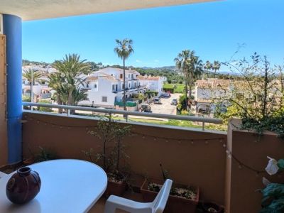 Balcon avec table et chaise, offrant des vues sur un quartier résidentiel avec maisons blanches, palmiers et collines verdoyantes sous un ciel bleu clair.