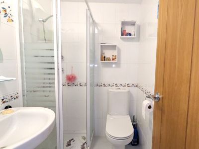 Salle de douche moderne avec murs carrelés blancs et frise décorative. Comprend une cabine de douche vitrée à bandes dépolies, un lavabo suspendu et des toilettes, avec deux étagères murales.