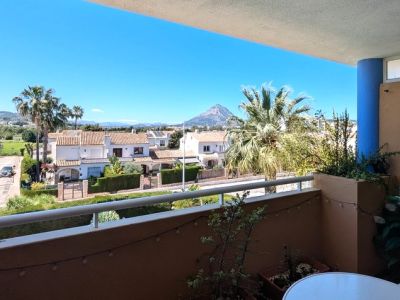 Terrasse ouverte avec balustrade blanche et plantes en pot, offrant des vues étendues sur les résidences locales, les zones vertes et le Montgó sous un ciel clair.