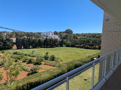 Vue depuis la terrasse privée avec balustrade moderne en verre et métal, donnant sur de vastes champs verts, des arbres et des bâtiments lointains sous un ciel clair.