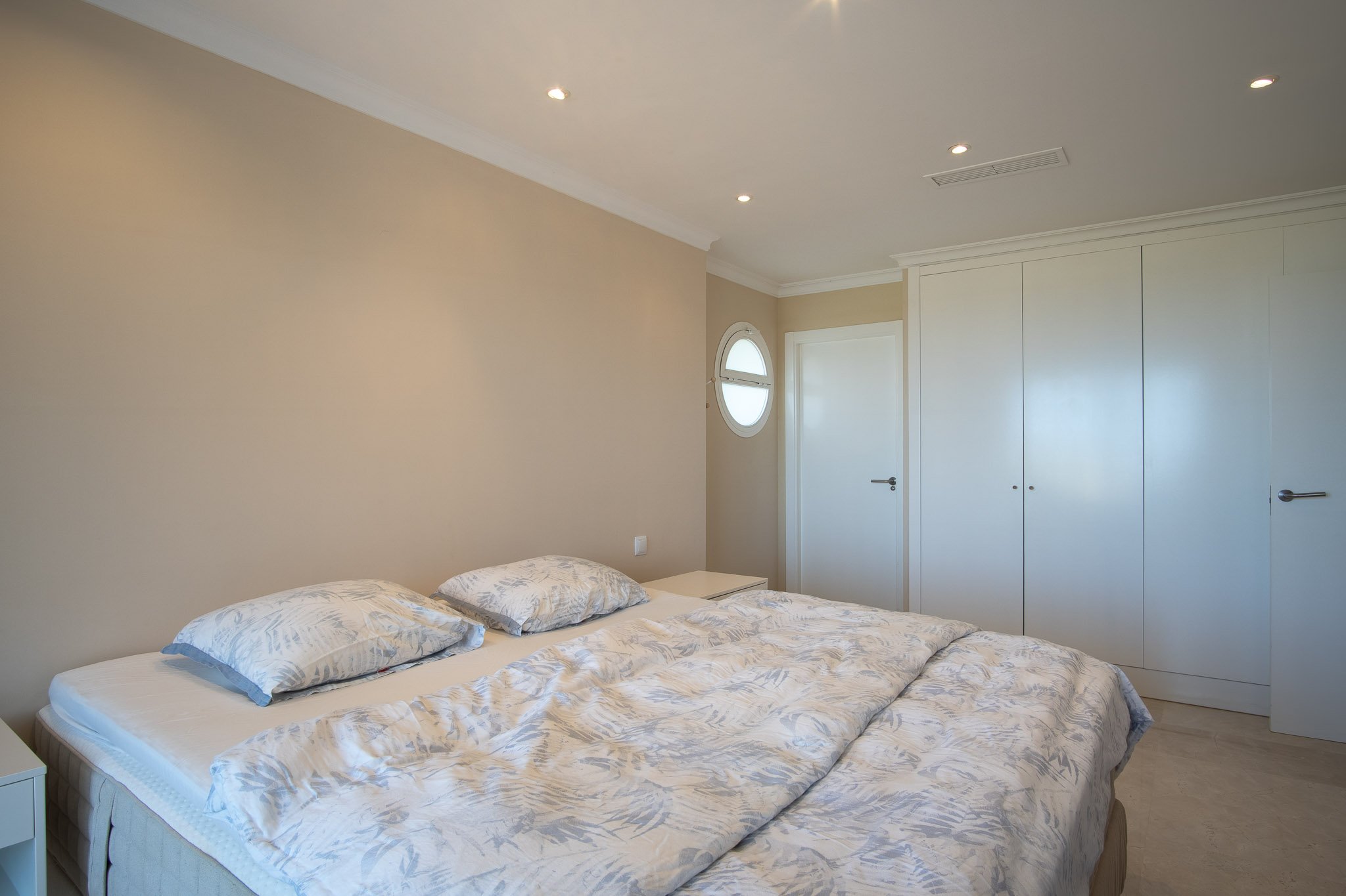 Chambre avec lit double, placards encastrés blancs et sol en marbre. La pièce dispose de spots encastrés au plafond et d'une fenêtre œil-de-bœuf.