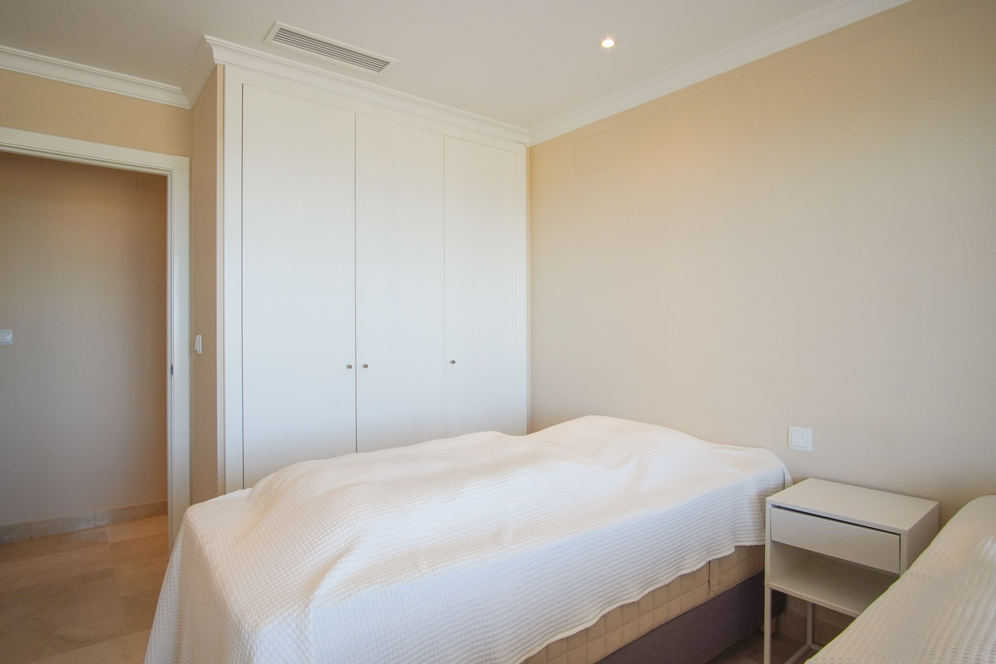 Chambre lumineuse avec placards intégrés blancs, sol en marbre et climatisation gainable. Murs aux tons crème et moulures de plafond classiques.